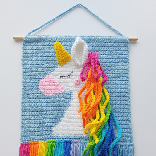Crocheted unicorn wall hanging with colourful mane and rainbow tail on a light blue background.
