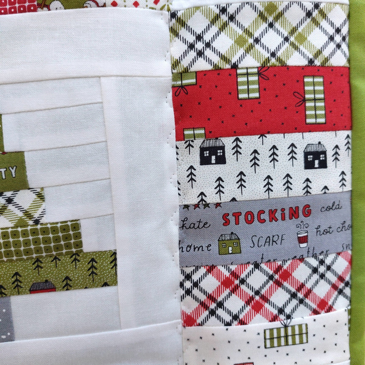 Close-up of a handmade quilt showing various fabric patterns including plaid, trees, houses, and a gray patch with the word 'Stocking'.