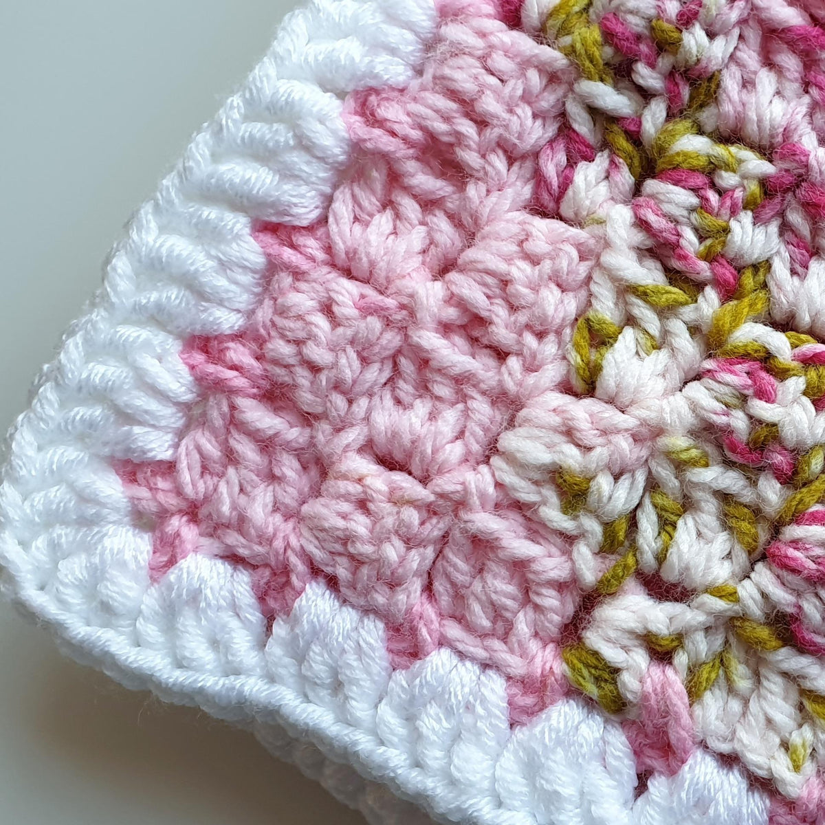 the corner of a crochet baby blanket, the corner is in white stitches and the other stitches are pink and green