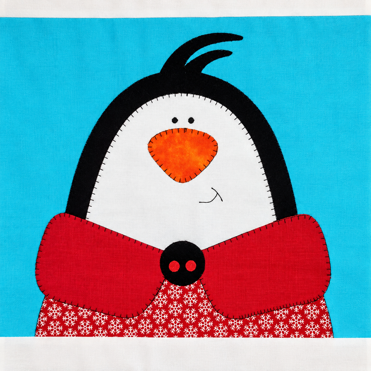 A stylized penguin illustration or textile artwork on a white border, with a bright blue background. The penguin has a white face, black outline, orange beak, and a red scarf with a red-and-white snowflake pattern.
