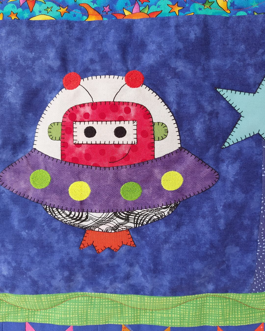 an alien spacecraft on a blue fabric background on an outer space quilt. The spacecraft has a smiling face in it.