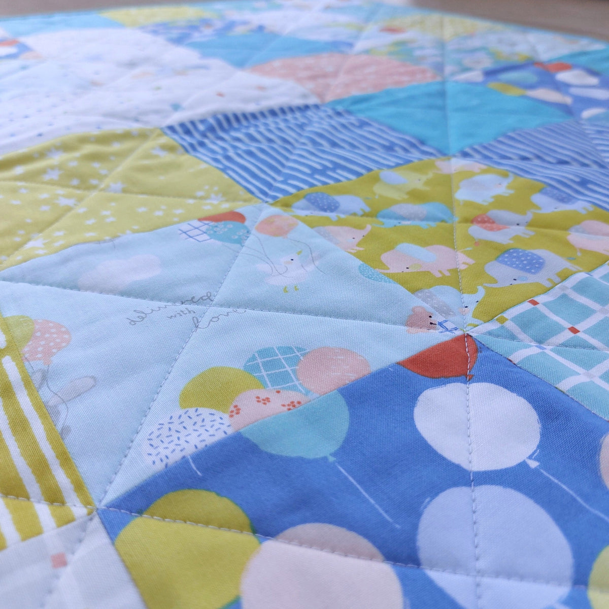 A close-up of a handmade patchwork quilt featuring various fabric panels with pastel colors, including blue, yellow, white, and pink, with balloon and cloud patterns.