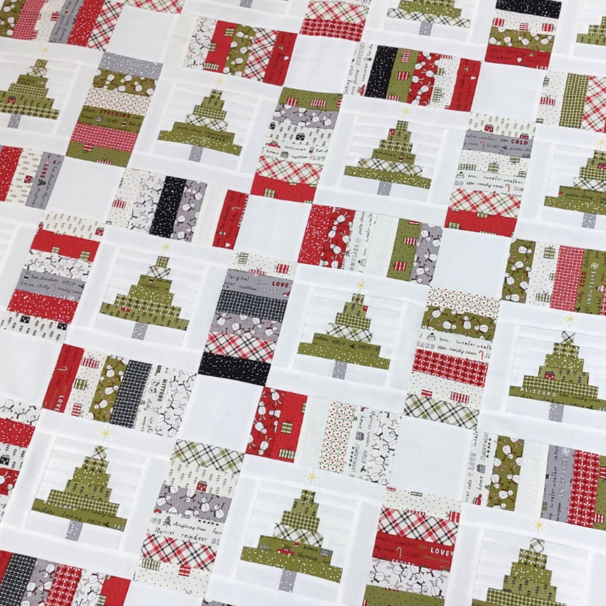 A close-up of a quilted fabric featuring a patchwork design of Christmas trees and holiday motifs in red, olive green, gray, black, and white.