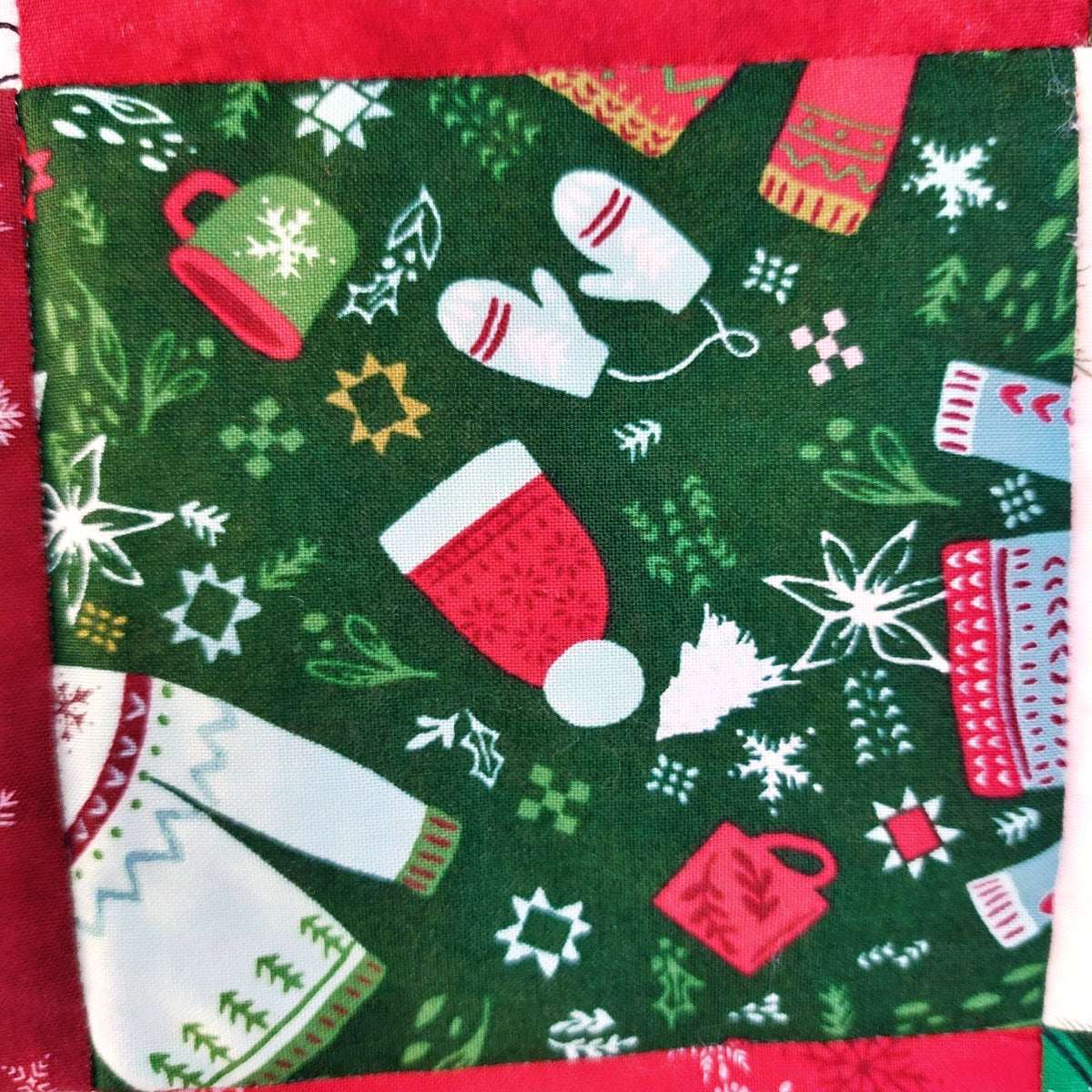 A close-up of a green fabric with a festive holiday print featuring red and white knit socks, white mittens, red mugs, white snowflakes, pine trees, and small green and yellow motifs, bordered by a red fabric edge.