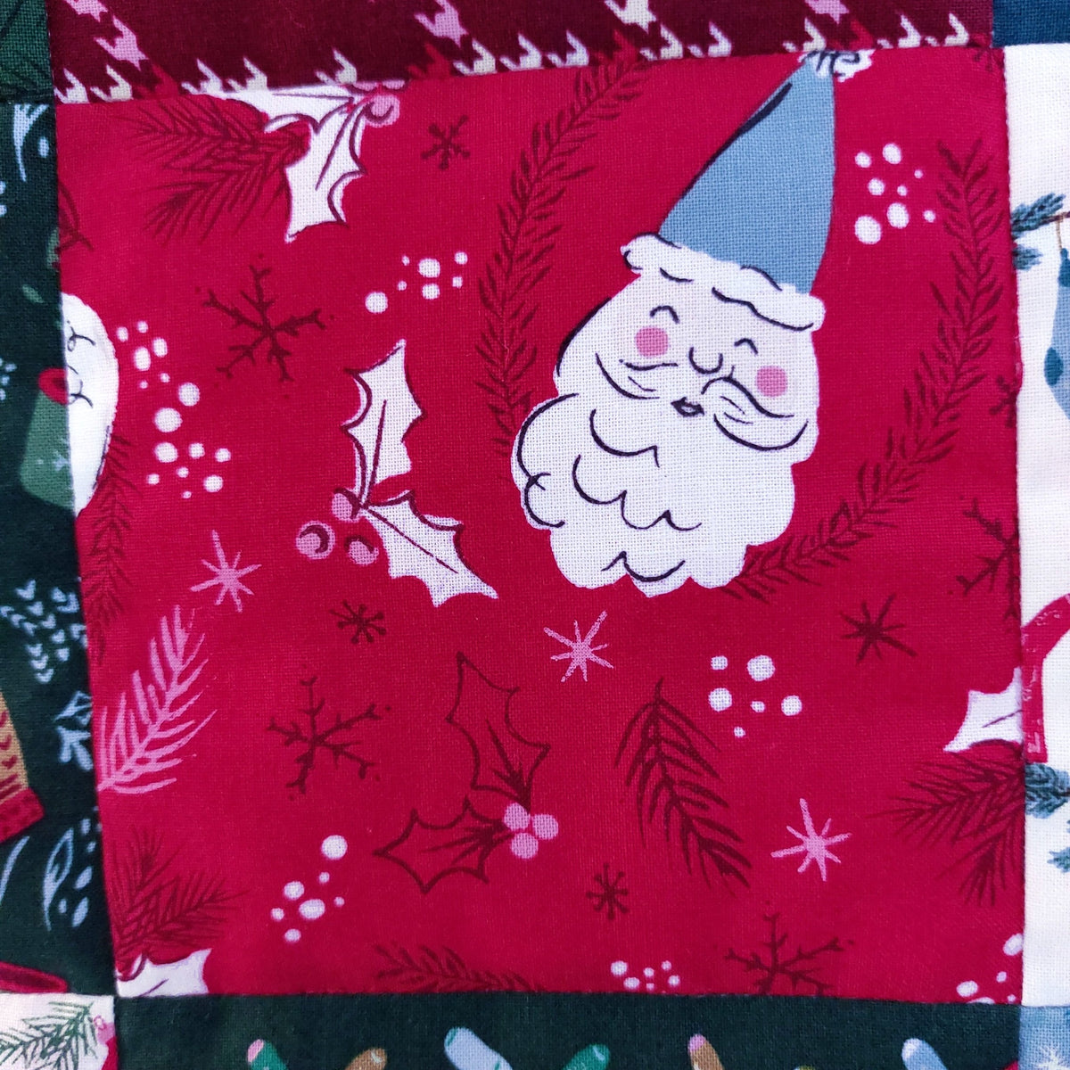 A close-up of a red Christmas-themed fabric featuring a white Santa face, blue hat, holly leaves, pine branches, snowflakes, and white berries, stitched with other patterned fabrics.