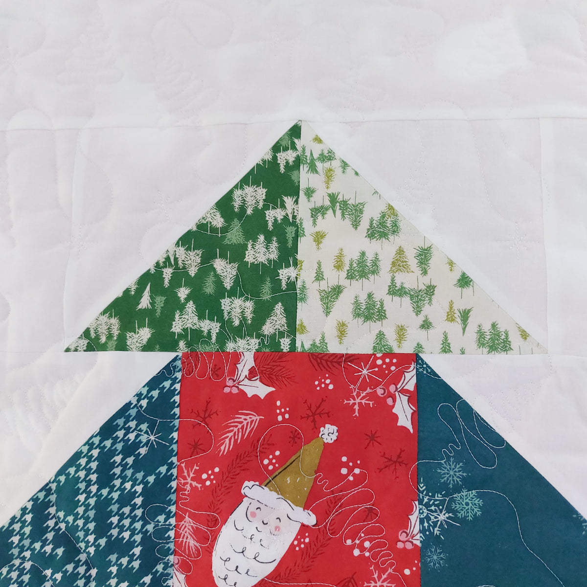 A close-up of a quilted fabric design featuring geometric triangle and square blocks with holiday patterns, including green and cream tree prints, a red Santa pattern, and teal houndstooth and snowflake designs on a white quilted background.