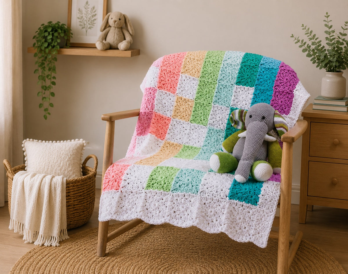 A colorful crocheted patchwork throw blanket draped over a wooden chair in a cozy, sunlit room. The blanket features a grid of pastel and bright crochet squares in pink, orange, yellow, green, blue, and purple on a white textured base.