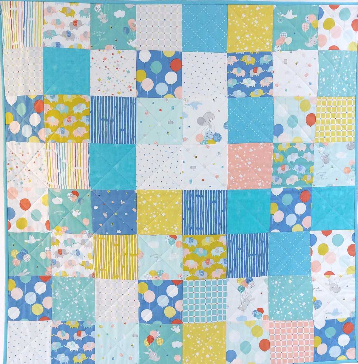 A square patchwork quilt featuring a grid of colorful fabric squares with various patterns including polka dots, stripes, florals, and clouds in shades of blue, teal, yellow, pink, and white.