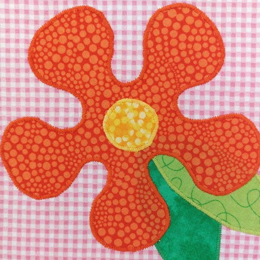 a pink check patchwork square with a bright orange flower which has a yellow centre and green leaves sewn on