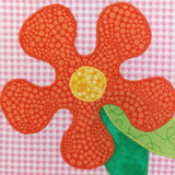 a pink check patchwork square with a bright orange flower which has a yellow centre and green leaves sewn on