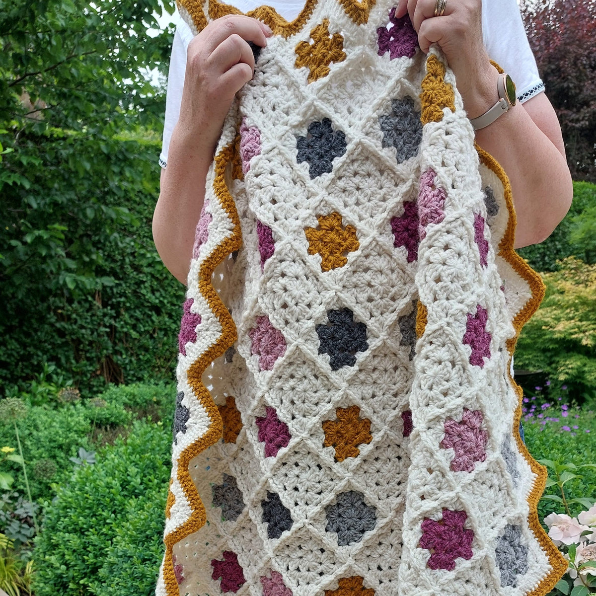 the handmade crochet granny blanket being held up with a garden backdrop showing the granny squares