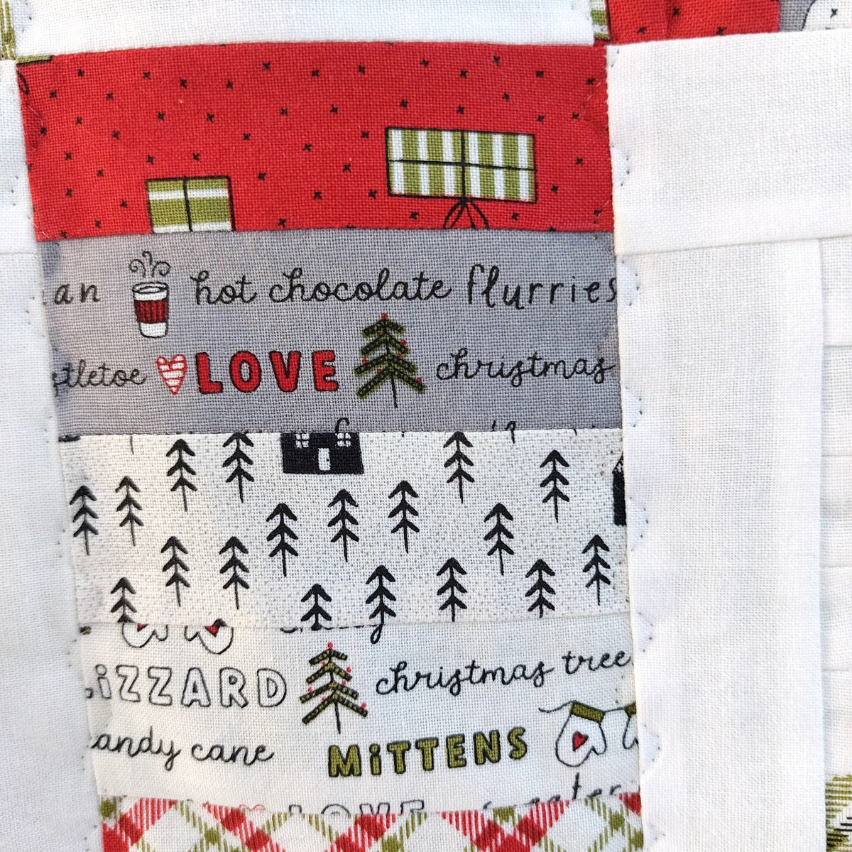 A close-up of a handmade quilt or fabric patchwork featuring various holiday-themed printed fabric strips in red, gray, white, and green.