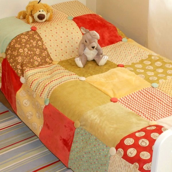 patchwork quilt on a bed. The quilt is made up of big squares of red green and yellow fleece with other squares of flowers.