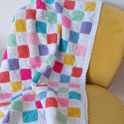 each rainbow coloured crochet square is next to an exact same square in white with the blanket draped on a yellow chair 