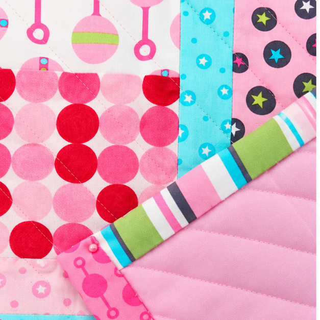 A close-up of a quilted fabric item, likely a baby blanket or quilt, showing multiple colorful patterned sections including pink polka dots, red circles, blue stars, black stars, and a striped border with pink, white, blue, green, and black.