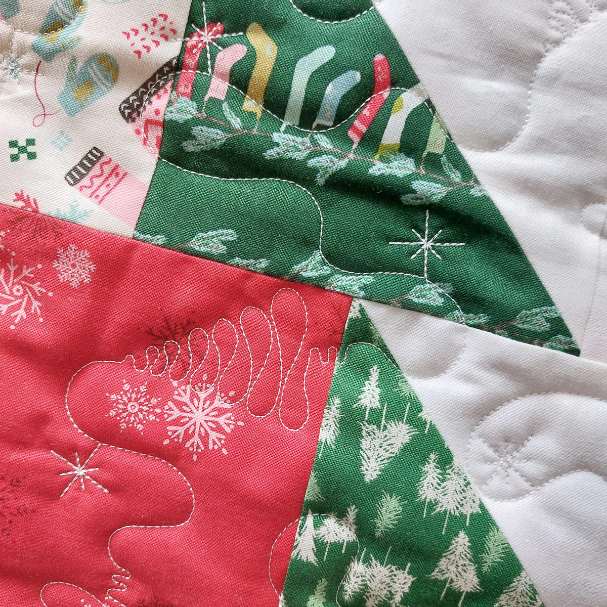 A close-up of a handmade quilted fabric patchwork featuring festive holiday designs, including a red section with white snowflakes, a green section with white pine trees, and a green section with white Christmas trees and snowflakes.