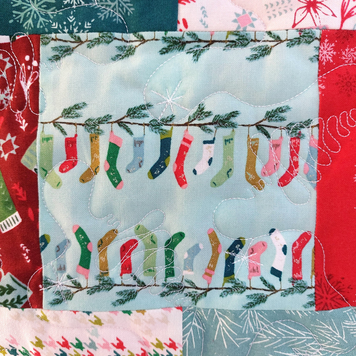 A close-up of a quilted fabric panel featuring a light blue center with embroidered Christmas stockings hanging from branches, surrounded by red, green, and white patterned quilted borders.
