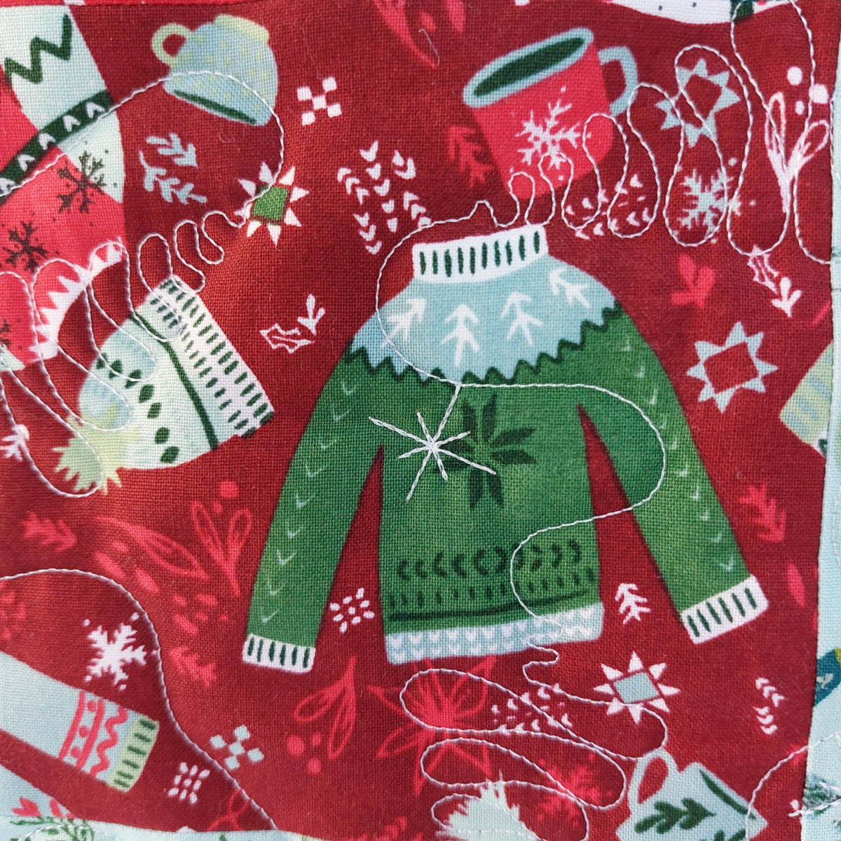 A close-up of a red fabric featuring a festive Christmas sweater and mug pattern, with a green sweater at the center and white stitched thread details.