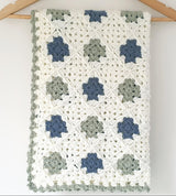 Handmade baby blanket hanging on a coat hanger. blanket has squares of blue green and cream