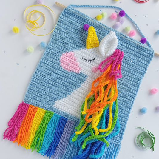 Crocheted unicorn wall hanging  with rainbow coloured hair and tassels on a light background