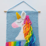 Crocheted unicorn wall hanging with colourful mane and rainbow tail on a light blue background.