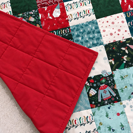 A close-up of a quilted red fabric panel laid diagonally over a colorful Christmas patchwork quilt with green, red, white, and blue holiday prints.
