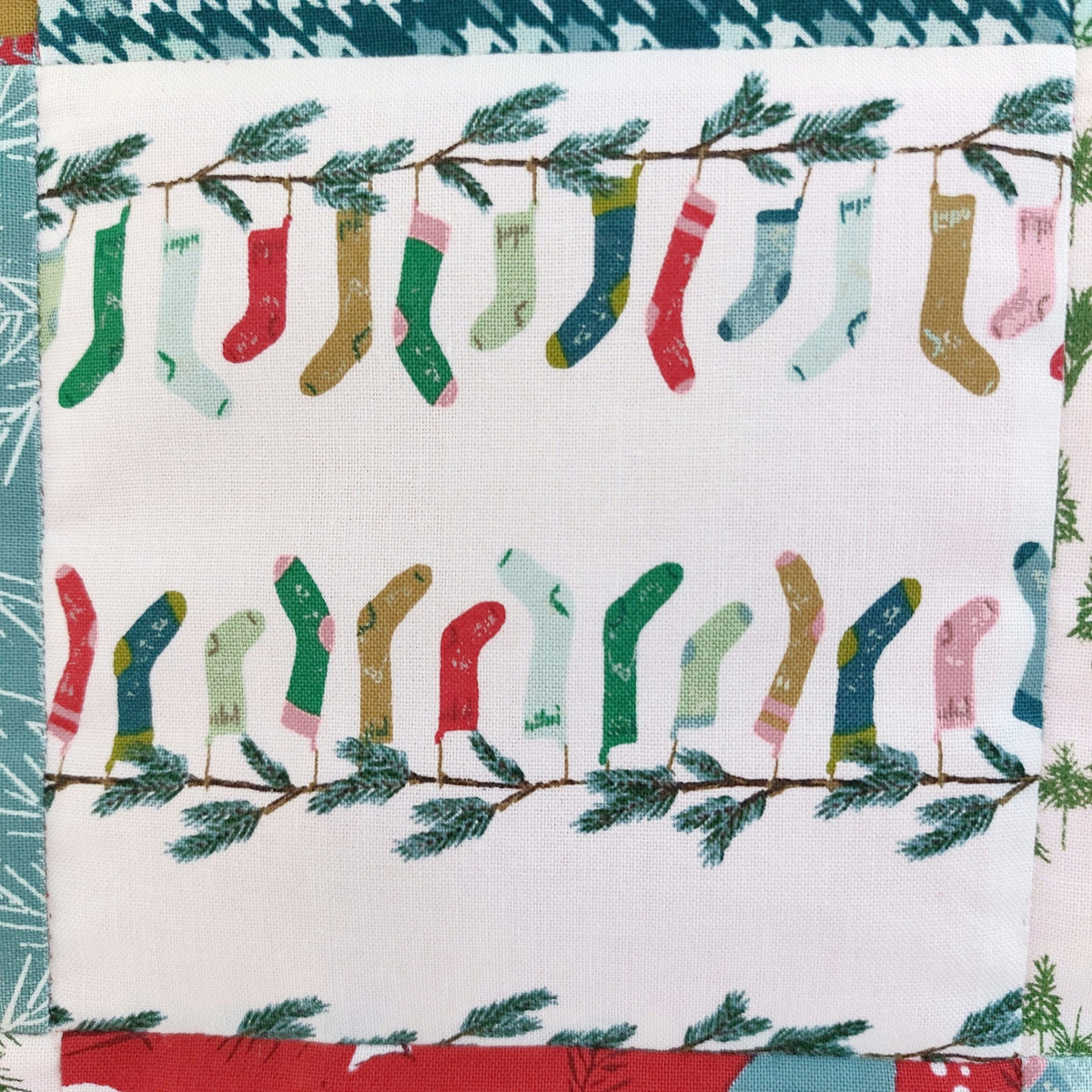 A close-up of a fabric product featuring a repeating pattern of colorful Christmas socks hanging from branches with green pine needles, on a white background.