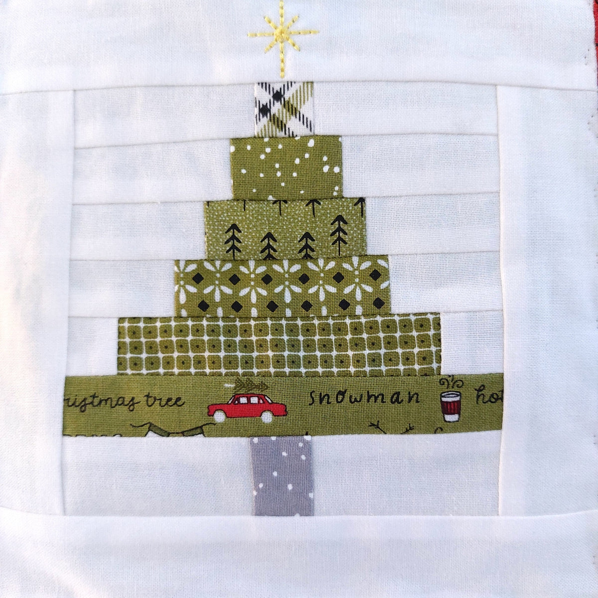 A close-up of a quilted Christmas tree block on a white fabric background. The tree is made of layered green patterned fabrics with a gold star on top, a gray trunk, and a green base with a red car and the words 'Christmas tree' and 'snowman'.