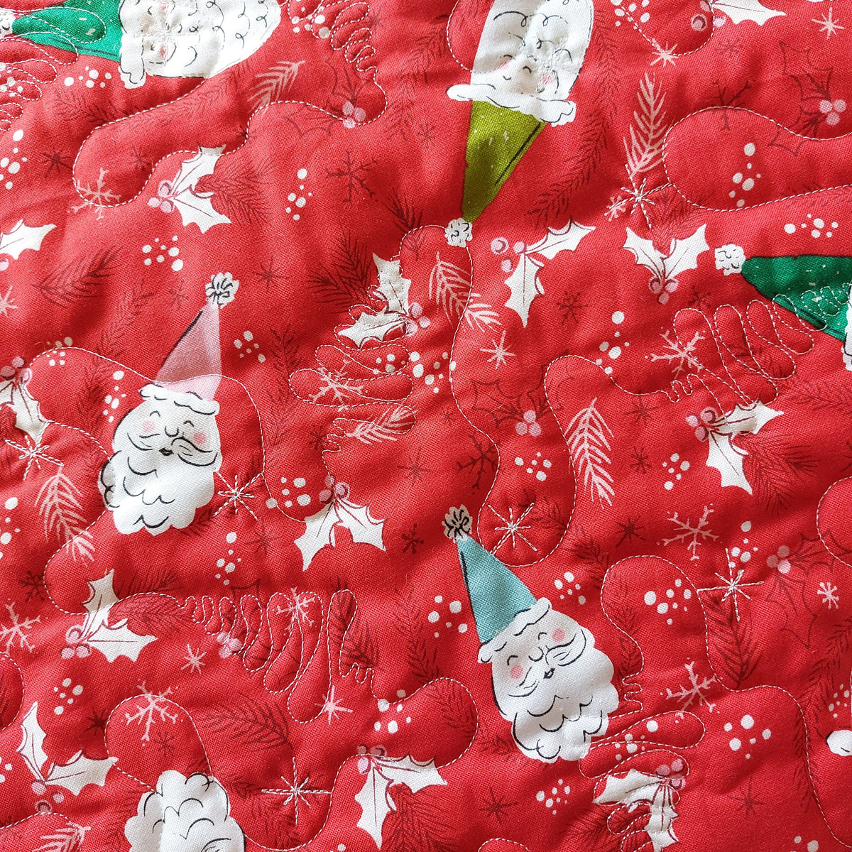 A close-up of a red quilted fabric featuring a festive Christmas pattern with embroidered Santa Claus faces, holly leaves, pine branches, snowflakes, and small white berries.