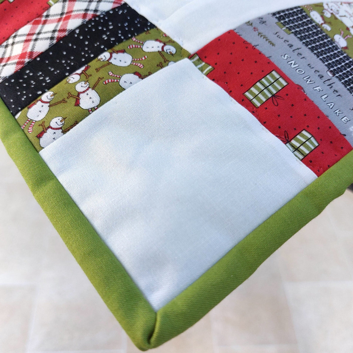 A close-up of a handmade quilted patchwork mat or cushion with a solid olive green fabric border and a multi-colored patchwork design featuring holiday-themed fabrics (snowman print, red plaid, black snow pattern, gray 'Snow Lake' print) and a large plain white fabric pocket.