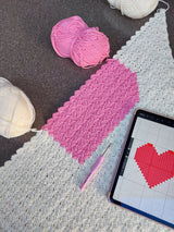 photo showing the handmade pink heart baby blanket being made with 3 balls of wool in cream and pink with a pattern on an i pad