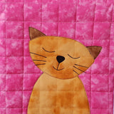 bright pink background with a gold act sewn on. The cat has dark brown ears, black whiskers and it's eyes are closed