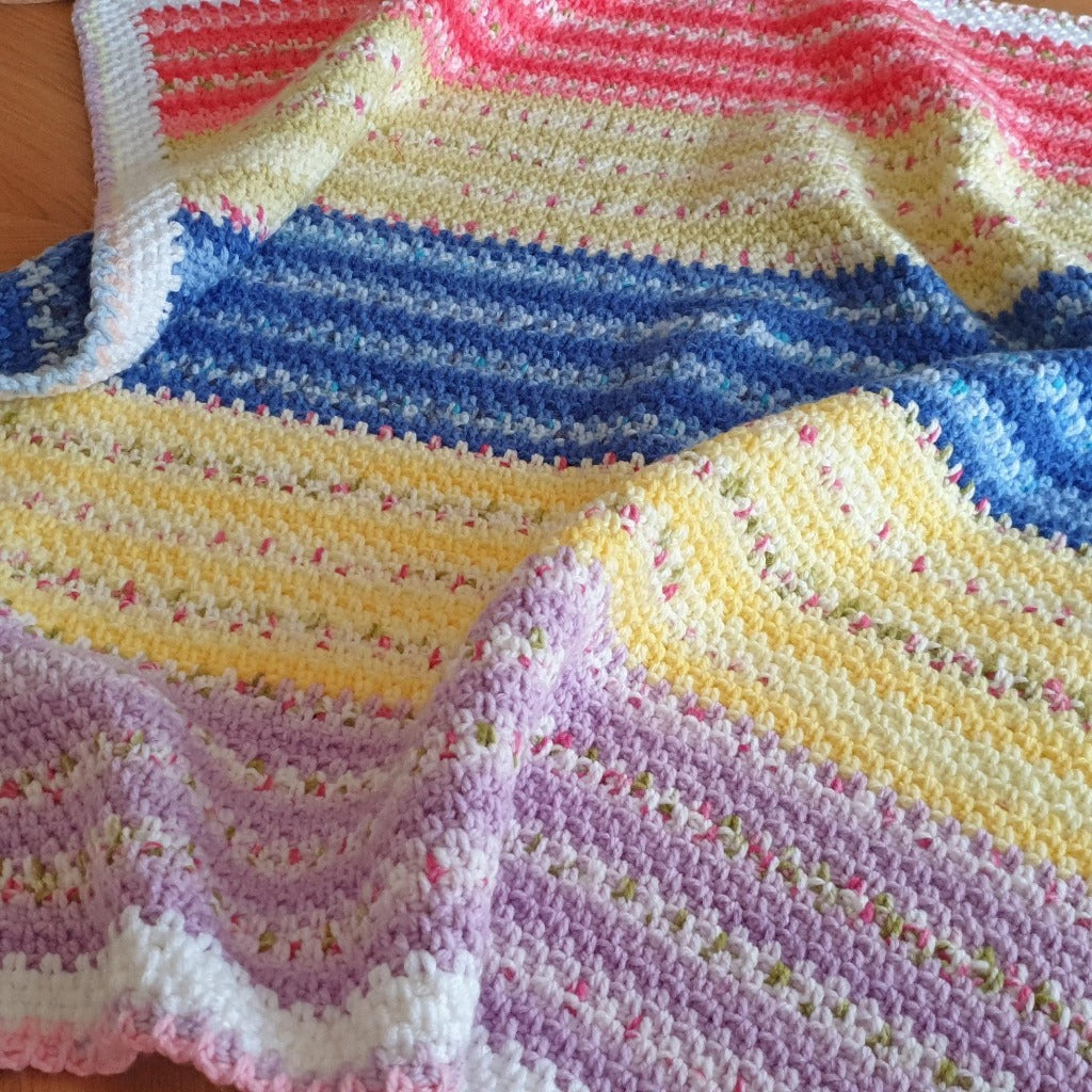 handmade baby blanket laid out showing the rows of stitches which change from purple to yellow to dark blue to green and then dark pink