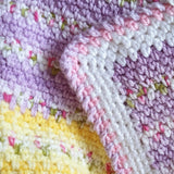 Handmade Babhandmade baby blanket showing the stitches of the blanket which change from yellow with pink and green flowers to lilac. It also show the corner of the baby blanket in white, pink and lilac