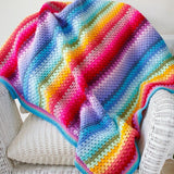 crochet blanket in stripes of pink, red, green, blue, purple and orange,. The blanket is draped on a white chair.