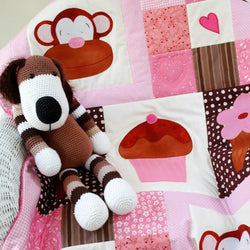 pink, white and brown patchwork squares with a monkey face on one, a large cup cake on one and an ice cream. There is a crocheted brown and white striped dog sitting on the quilt.