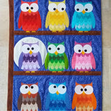 a quilt of a brown tree with three brown branches. On each branch are 3 owls in different colours of red, yellow, blue, pink, green and brown. On the middle branch one owl sits in front of a big white moon