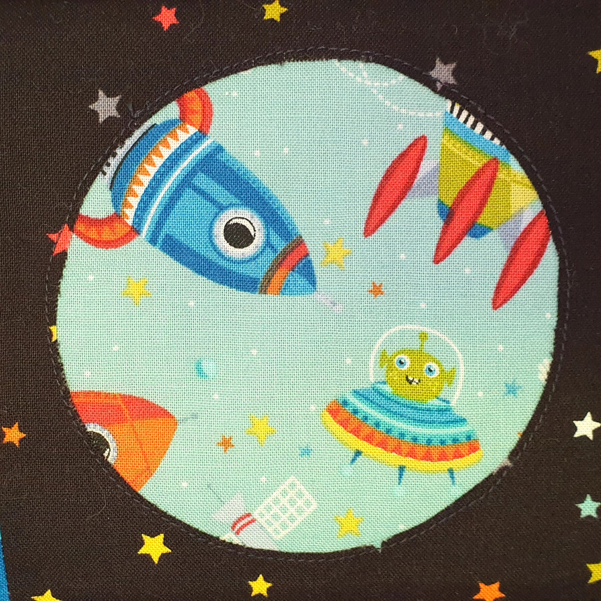 a black cotton background with a circle cut out which shows a blue rocket and an alien in a spaceship