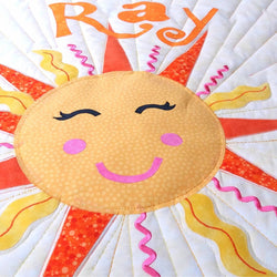 A large sun with a smiling face ob it with large, black eyelashes, pink cheeks and a pink smile. It has orange, yellow and pink flames coming from behind the sun 