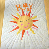 The whole quilt showing a smiling sun with large sun rays and the name of the baby at the top